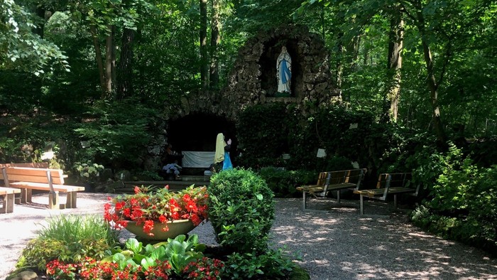 Ein kleiner Schrein mit einer Marienstatue in einem grünen Garten mit Bänken und Blumenbeeten.
