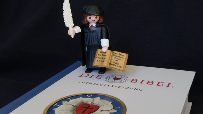 Eine Playmobil-Figur hält ein Buch und eine Feder über einem Buch mit dem Titel 