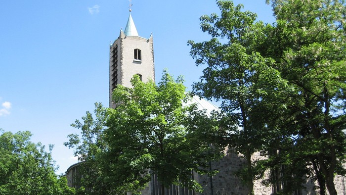 Historische Steinkirche mit hohem Glockenturm, eingerahmt von üppigen grünen Bäumen unter einem klaren blauen Himmel.