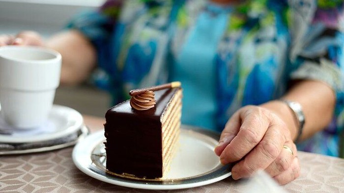 Eine Person hält ein Stück Schokoladenkuchen auf einem Teller, daneben steht eine Tasse Kaffee