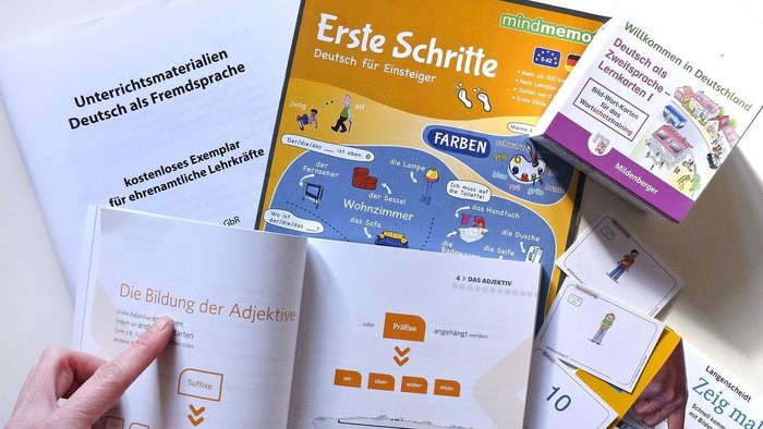 Eine Hand hält ein Buch mit dem Titel "Ersie Schrite", umgeben von verschiedenen Lernmaterialien und Karten.