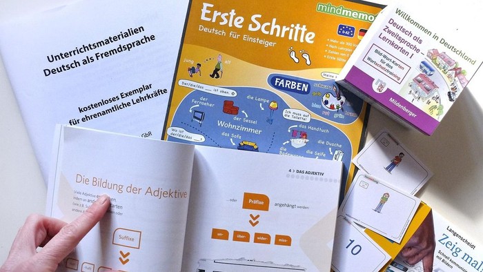 Eine Hand hält ein Buch mit dem Titel "Ersie Schrite", umgeben von verschiedenen Lernmaterialien und Karten.