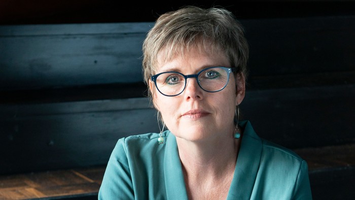 Kvinde i blå bluse og briller sidder på benk.
