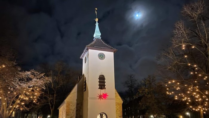 Nachtansicht einer Kirche mit einem Uhrturm, beleuchtet von Lichtern und einem Vollmond am Himmel.
