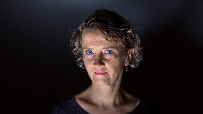 En kvinde med kort, krøllet hår og blå øjne ser alvorligt ud.