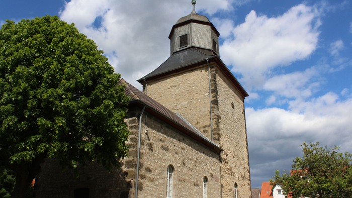 Steinerne Kirche mit hohem Turm und Uhr, umgeben von Bäumen unter blauem Himmel.