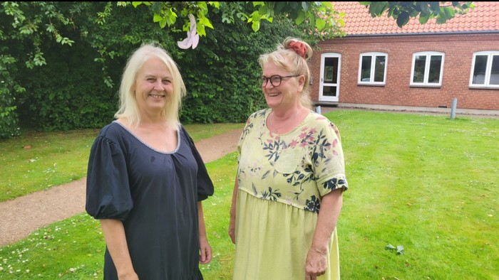 To dyrkede kvinder står i en have foran et rødt hus.