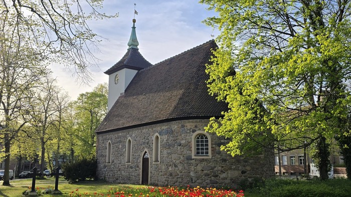 Kleine Kirche mit spitzem Turm, umgeben von Bäumen und blühenden Blumen