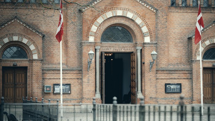 Bygningen har to danske flag og en central indgang med bueformet vindue.