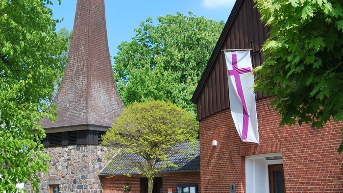 Das Bild zeigt ein Ziegelgebäude mit einem hohen Schornstein und einem Kreuzbanner.