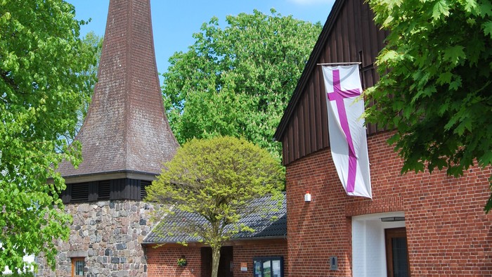 Das Bild zeigt ein Ziegelgebäude mit einem hohen Schornstein und einem Kreuzbanner.