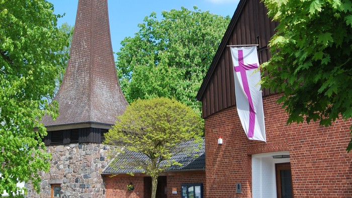 Das Bild zeigt ein Ziegelgebäude mit einem hohen Schornstein und einem Kreuzbanner.
