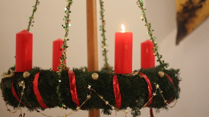 Ein Adventskranz mit vier roten Kerzen, geschmückt mit grünem Tannengrün und goldenen Dekorationen, hängt an einem Holzpfahl