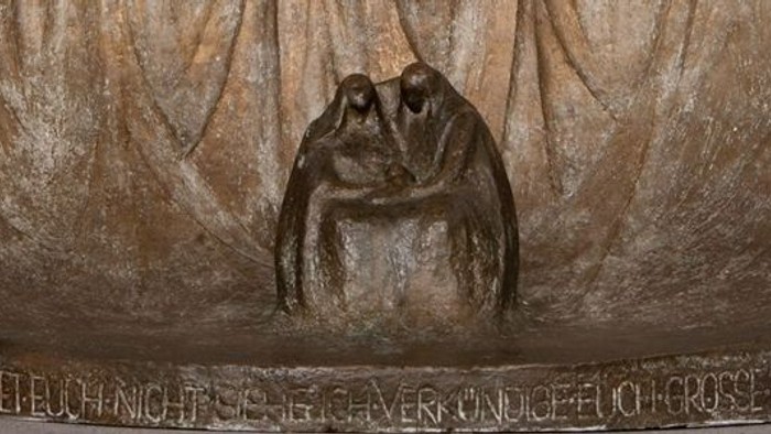 Das Bild zeigt eine Bronzestatue von zwei Menschen, die sich umarmen, vor einem texturierten Hintergrund.