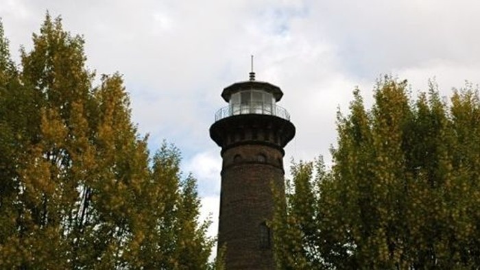 Ein hoher Leuchtturm steht zwischen Bäumen mit herbstlicher Blattfärbung unter einem bewölkten Himmel.
