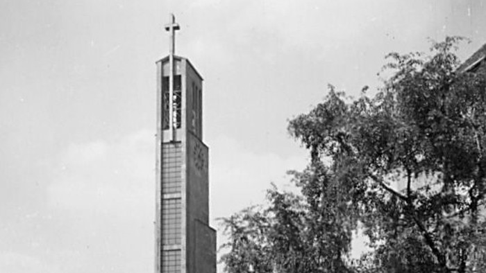 Kirche mit hohem Turm und Kreuz, alter Wagen in der Nähe geparkt, im Hintergrund Bäume und Gebäude.