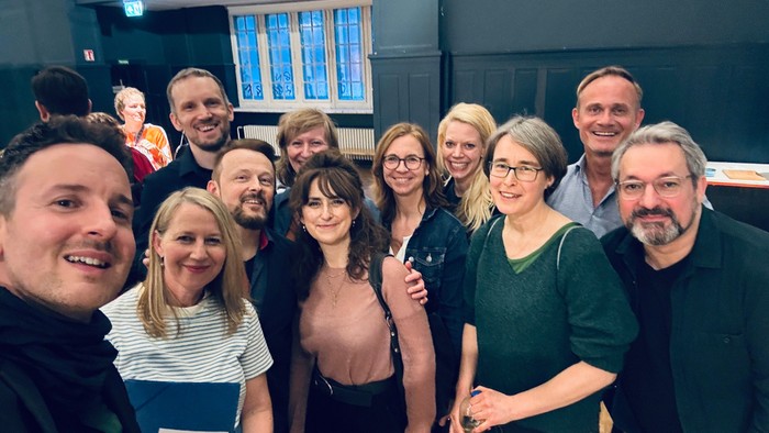 Eine Gruppe von Menschen posiert für ein Selfie in einem Raum mit großen Fenstern.