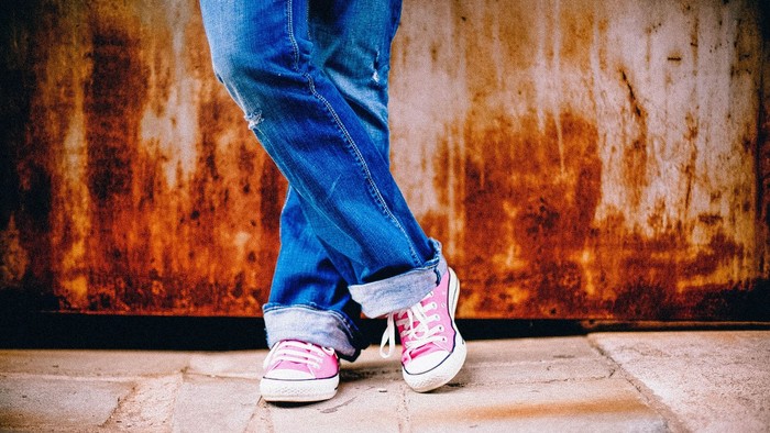 Person in blauen Jeans mit pinken Turnschuhen steht lässig vor einer rostfarbenen Wand