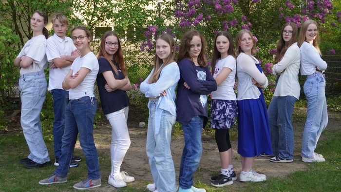 Eine Gruppe Jugendlicher posiert selbstbewusst im Freien mit violetten blühenden Sträuchern im Hintergrund.