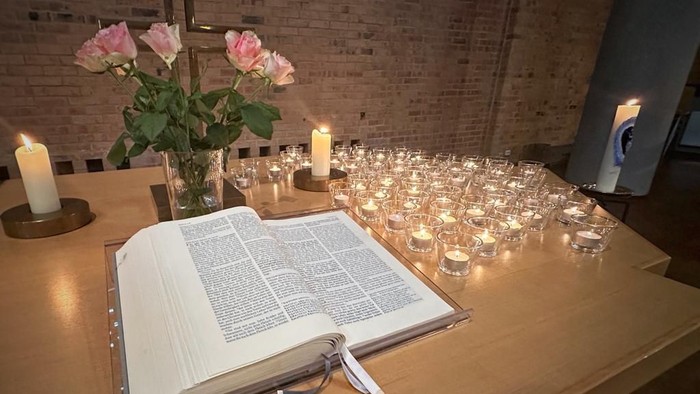 Elegantes Holztisch-Set mit offener Bibel, Kerzen, Blumengesteck und brennenden Votivkerzen in einer ruhigen Atmosphäre.