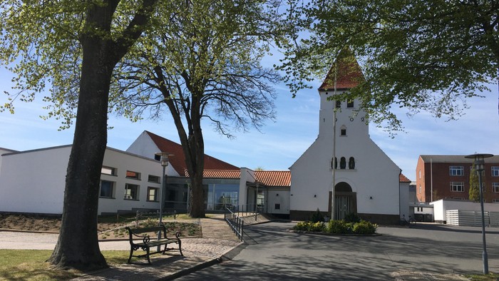 Et hvidt kirkebygning med spiret og røde tag, omgivet af træer og en bænk.