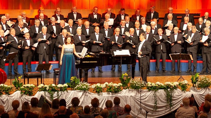 Ein großer Chor und Orchester treten in einem Konzert auf, begleitet von einem Dirigenten.