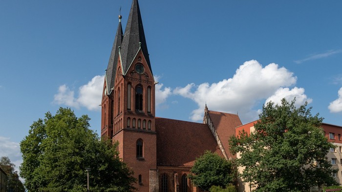 Kirchengebäude mit hohem Turm und spitzem Dach, umgeben von Bäumen und Grasfläche