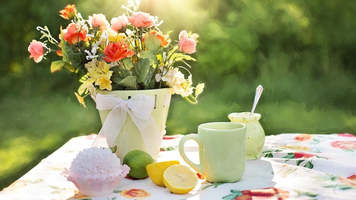 Helles Blumenarrangement in einem Eimer mit Zitronenscheiben, Krug und Tassen auf einem geblümten Tischtuch im Freien.