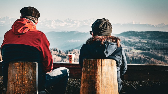Zwei Personen sitzen auf Holzstümpfen und genießen gemeinsam eine atemberaubende Berglandschaft.