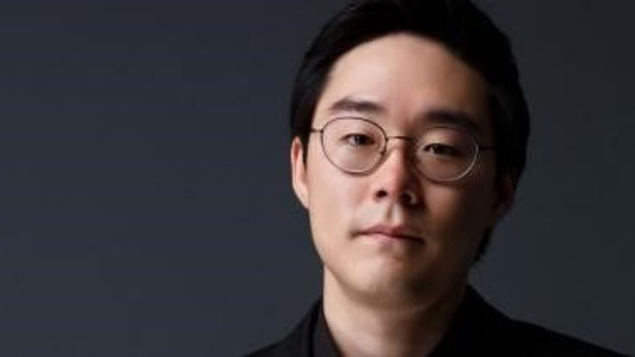 Ein Mann mit Brille und dunkler Kleidung vor einem grauen Hintergrund.