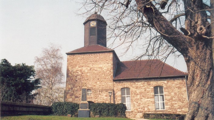 Kleine Steinkirche mit rotem Dach und Uhrturm, umgeben von Bäumen und Gras.