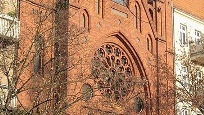 Gothische Backsteinkirche mit filigranem Fenster und Türbogen, umgeben von Bäumen und anderen Gebäuden.