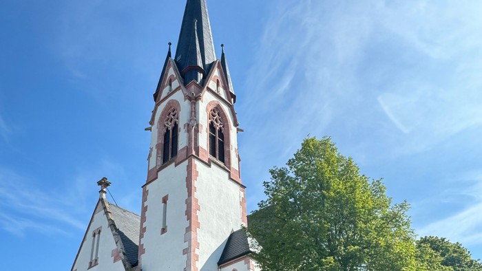 Weiße Kirche mit hohem Turm und spitzen Dach, umgeben von Bäumen und einem klaren blauen Himmel.