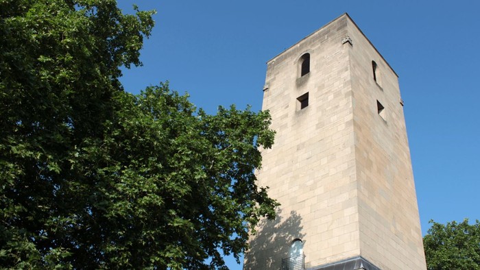 Ein hoher, weißer Turm ragt hinter Bäumen in den klaren blauen Himmel.