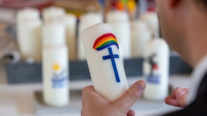 Hand hält Kerze mit Regenbogen und Kreuz vor mehreren Kerzen