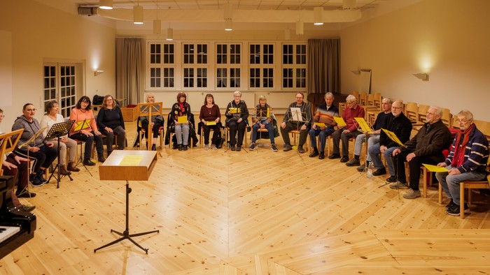 Eine Gruppe von Menschen sitzt in einem Kreis auf Stühlen in einem Raum mit Holzboden und großen Fenstern.