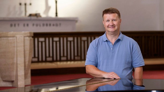 En mand i en blå polo-trøje sidder ved et klaver inde i en kirke med religiøse dekorationer.