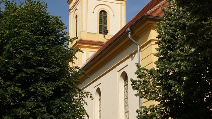 Kleine Kirche mit hohem Turm und Uhr, umgeben von Bäumen