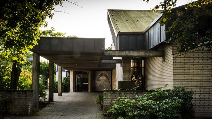 Das Bild zeigt ein modernes Gebäude mit Flachdach und Säulengang, umgeben von Grünpflanzen.