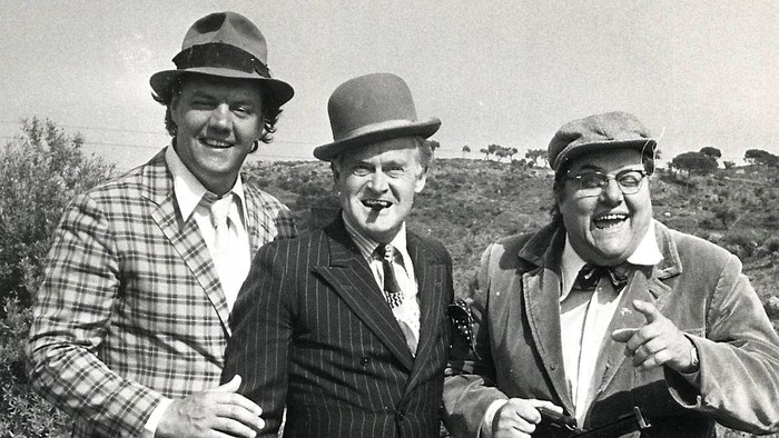 Tre mænd i vintage hatte og jakkesæt poser muntert udendørs og udstråler klassisk komisk charme.