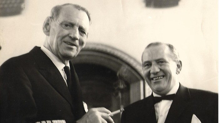 To formelt klædte mænd, der smiler og giver hånd udendørs på et vintage sort-hvidt fotografi.