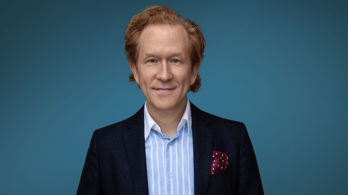 Professionel mand i mørk blazer, lyseblå skjorte og lommetørklæde, der smiler selvsikkert.