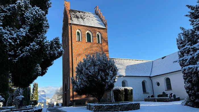 En snede kirke med tårn og hvide tag i snefaldende landskab