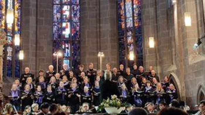 Eine große Gruppe von Menschen in einem prächtigen Saal, einige stehend, andere sitzend, mit bunten Glasfenstern im Hintergrund.
