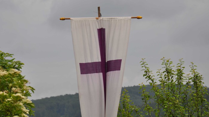 Weiße Flagge mit violettem Kreuz an einer Stange vor bewölktem Himmel.