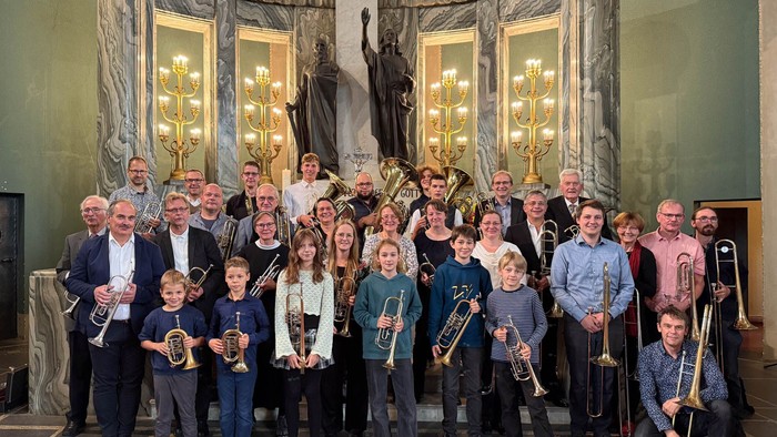 Eine Gruppe von Musiker:innen, darunter Erwachsene und Kinder, posiert gemeinsam mit Blechblasinstrumenten in einem prunkvollen Innenraum.