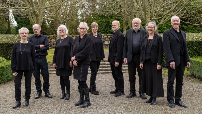 En gruppe af ni personer står sammen i en have, alle klædt i sort.