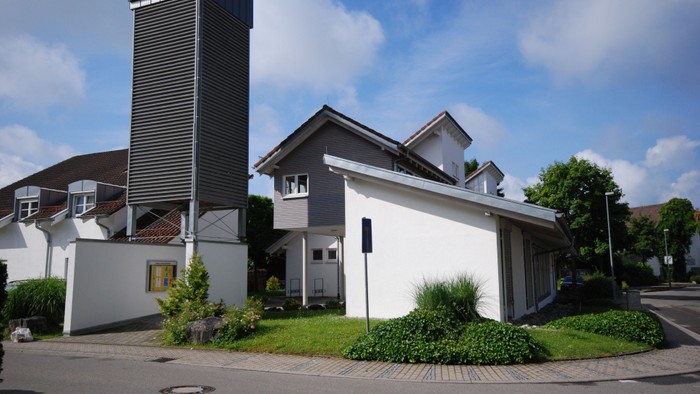 Ein modernes Gebäude mit hohem Kamin und weißer Fassade, umgeben von grünem Rasen und Bäumen.