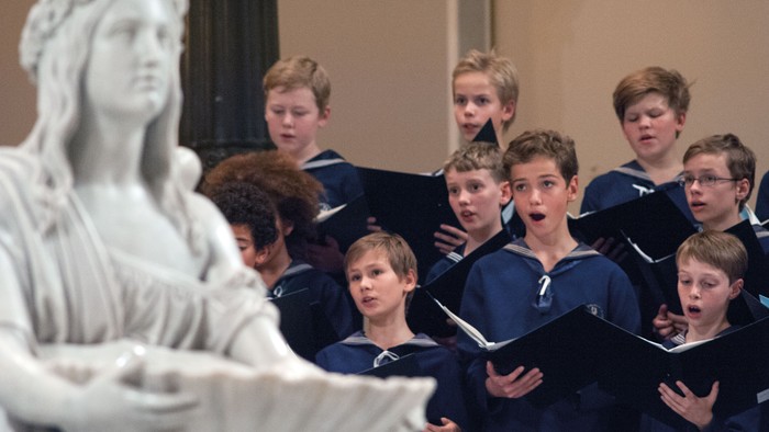 En gruppe drenge i mørke blå uniformer synger i en kirke, mens en statue af Jesus ser til.