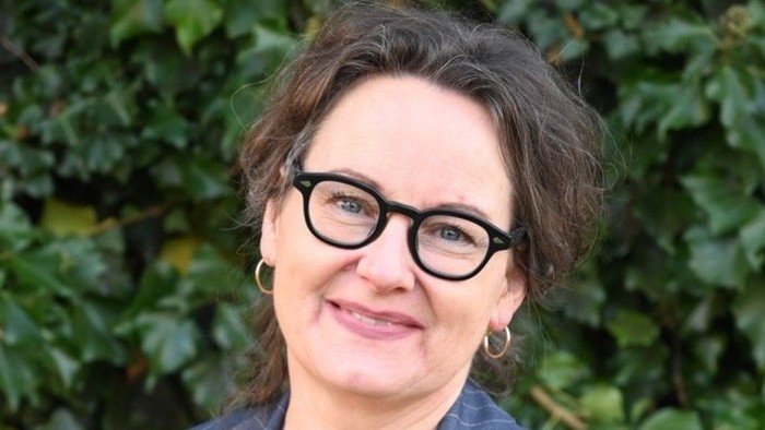 En kvinde med briller og kort hår, der smiler og står foran et grønt plantebak.